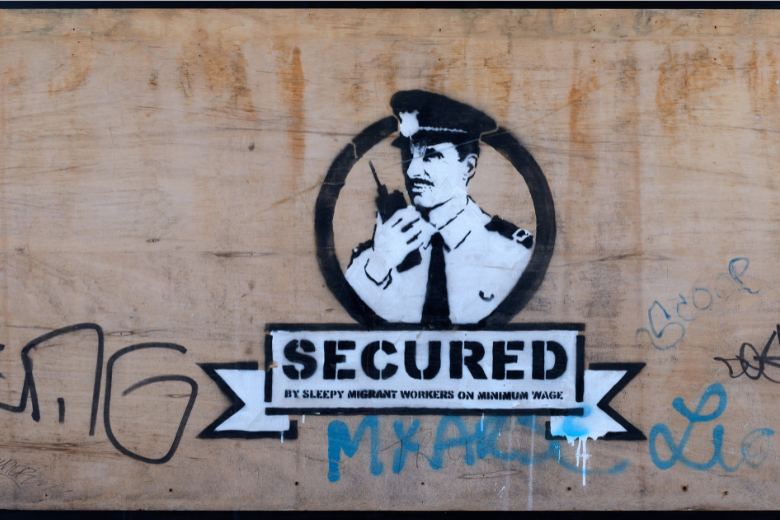 Banksy artwork titled 'Secured' displayed on a wooden surface, highlighting social commentary on migrant workers.