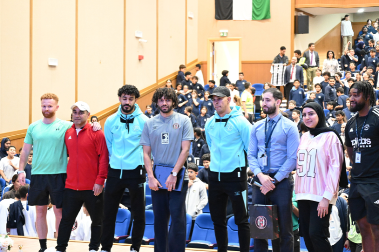 Mohamed Elneny and Al Jazira Club team members stand with ICS Al Danah staff on stage during a packed assembly.