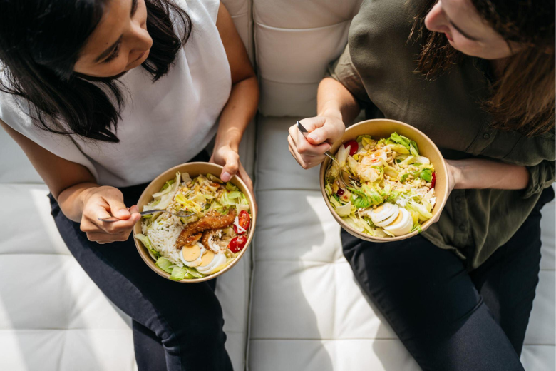 Two women enjoying healthy protein-packed lunch bowls from Meals on Me while seated together.