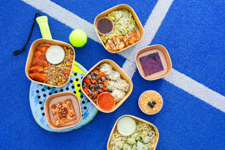High protein meal prep boxes from Meals on Me placed on a tennis court with healthy mains, sides, and desserts.