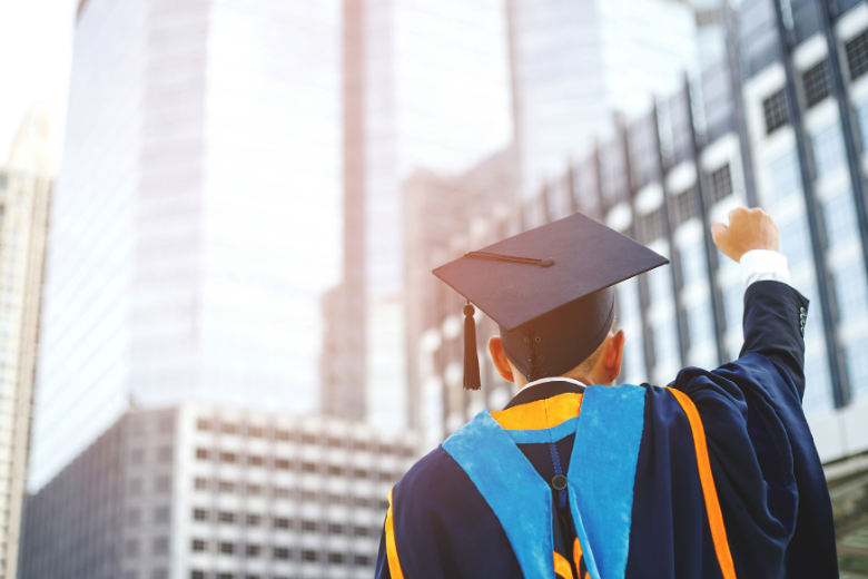 University graduate raising a fist in celebration in front of modern city buildings.